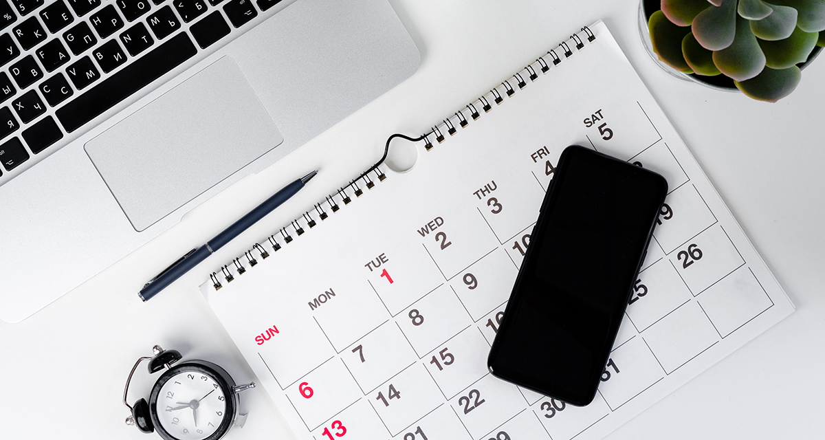 A flat-lay aerial view of a desk workspace featuring a spiral-bound wall calendar open to a month beginning on Tuesday the 1st, surrounded by a laptop keyboard, a black ballpoint pen, a small analog alarm clock, a black smartphone, and a small green succulent plant, all arranged on a white surface.