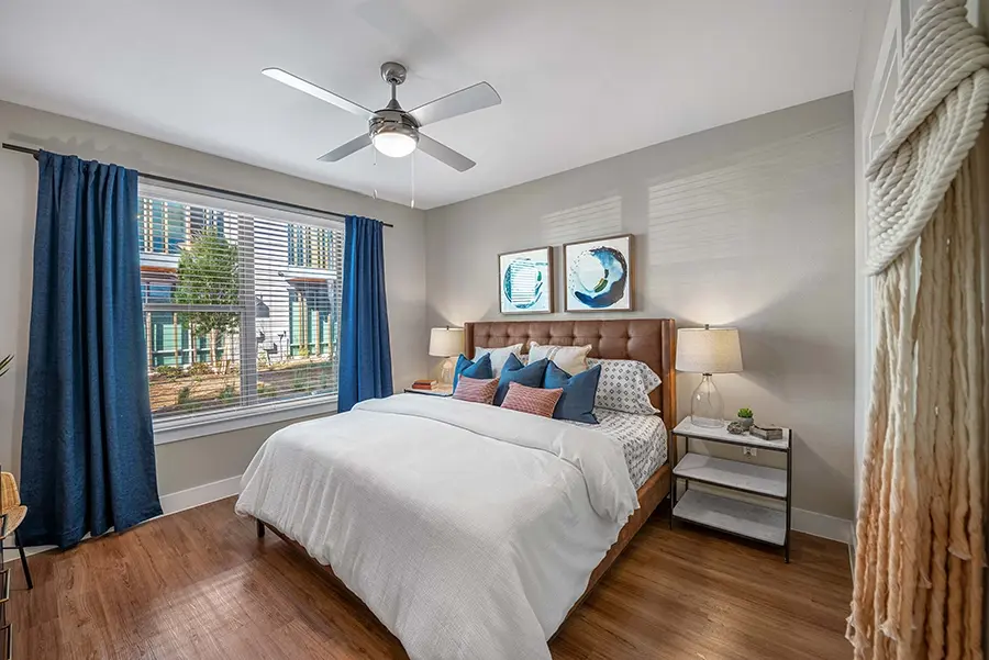 A furnished apartment bedroom with wood floors, white bedding, and blue curtains. 