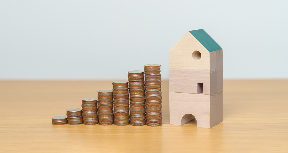 Stacks of coins increasing in height beside a small wooden house on a table, symbolizing rising housing costs, rent growth, or increased property value in the housing market.