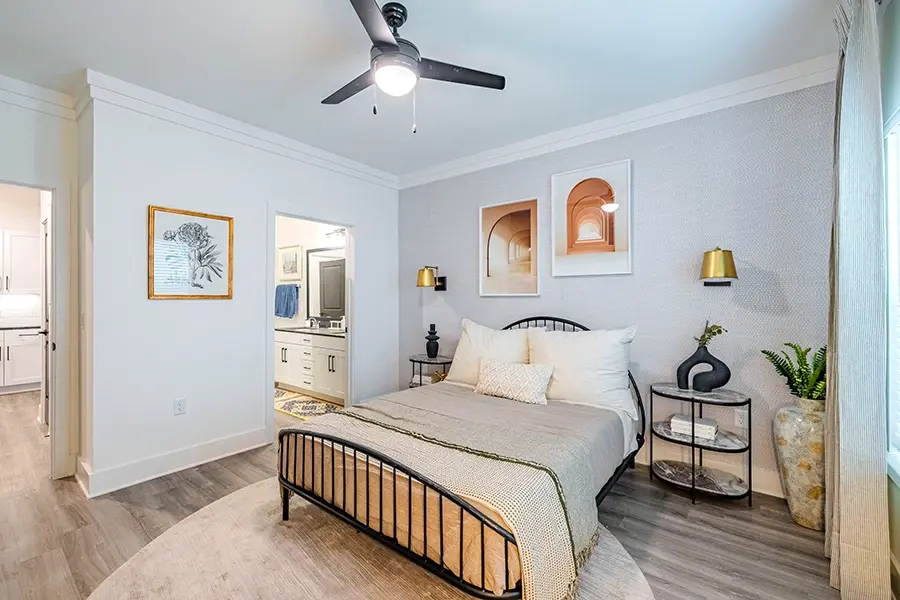 A furnished apartment bedroom with tan bedding, a round rug, and a view into the bathroom and hallway.