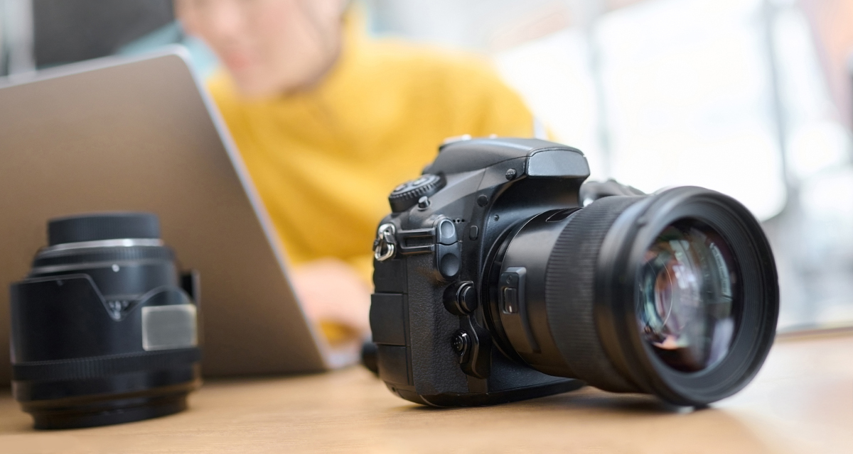 Close-up of a professional DSLR camera with a large lens placed on a wooden table next to an extra camera lens. In the blurred background, a person wearing a yellow sweater works on a laptop, suggesting a photography or editing workspace.