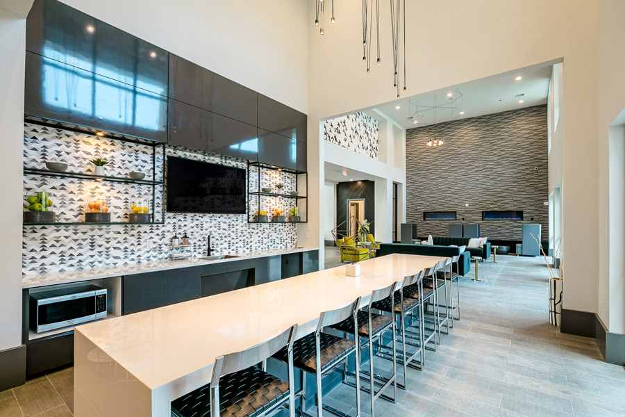 Long communal bar with light wood countertop, row of stools, and a dark backsplash with screens above.