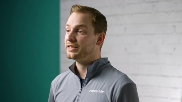 Smiling RentVision team member in a gray branded quarter-zip standing in front of a white brick wall in a bright office.