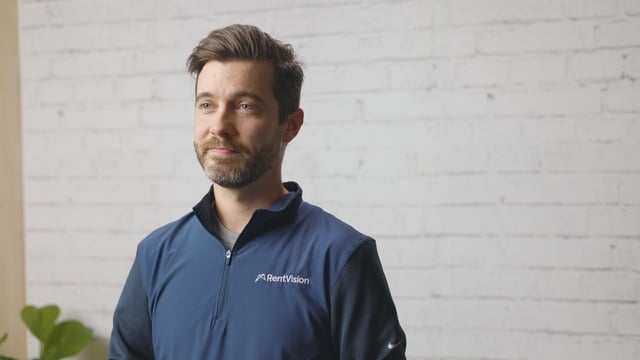 Smiling RentVision team member in a navy branded quarter-zip standing in front of a white brick wall in a bright office.