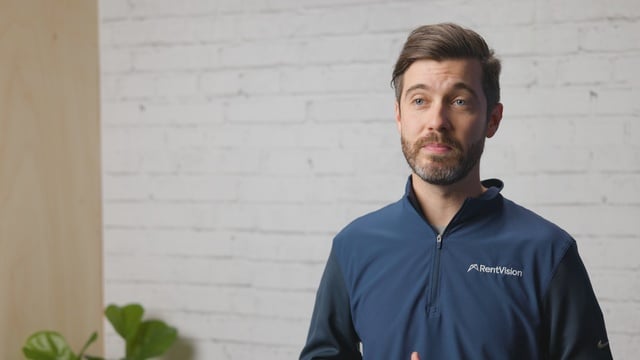Smiling RentVision team member in a navy branded quarter-zip standing in front of a white brick wall in a bright office.