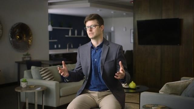 Man in glasses and a gray blazer speaking and gesturing during an interview in a modern apartment community lounge.