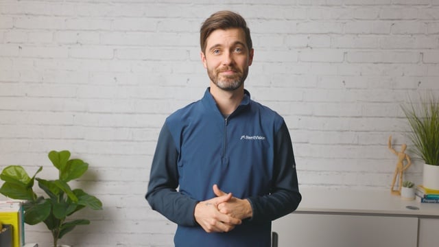 Smiling RentVision team member in a navy branded quarter-zip standing in front of a white brick wall in a bright office.