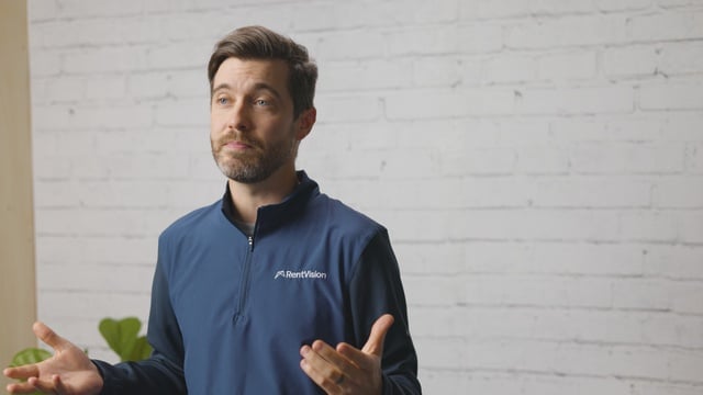 Smiling RentVision team member in a navy branded quarter-zip standing in front of a white brick wall in a bright office.
