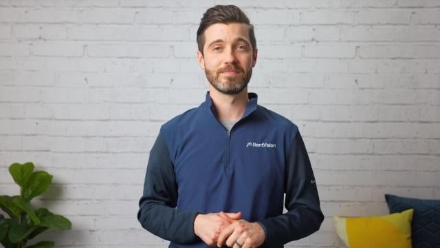 Smiling RentVision team member in a navy branded quarter-zip standing in front of a white brick wall in a bright office.