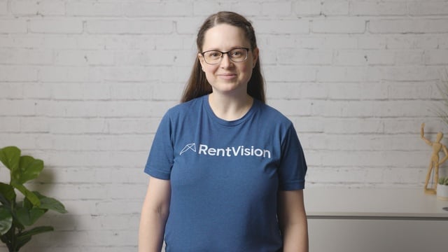 Smiling RentVision team member in a navy branded quarter-zip standing in front of a white brick wall in a bright office.