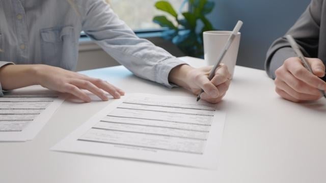 Two people reviewing and filling out paperwork together at a white desk, one signing a form with a pen.