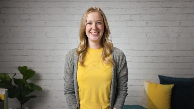 Smiling RentVision team member in a yellow branded t-shirt standing in front of a white brick wall in a bright office.